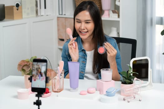 Young beautiful Asian reviewing cosmetic makeup brush cheek testing collection with pastel color, showing glow blusher mood with pink to promote on social media online recording smartphone. Stratagem.