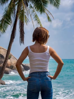 A young girl in jeans and a white T-shirt stands and looks at the sea under a palm tree
