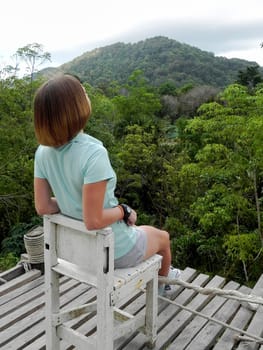 Woman hiker sitting alone on a wooden chair overlooking the green mountains. View point on Koh Phangan