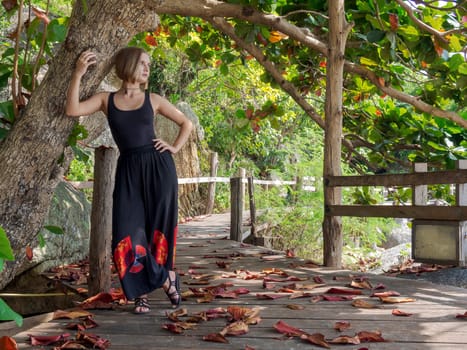 A young woman in black clothes poses by a tree in a rainforest on a wooden floor in fallen red foliage. walk in the forest on a tropical island