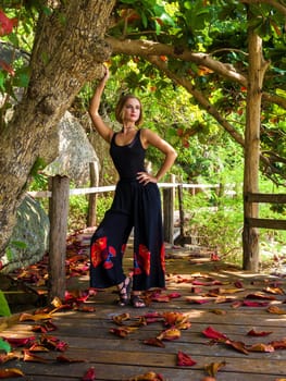 A young woman in black clothes poses by a tree in a rainforest on a wooden floor in fallen red foliage. walk in the forest on a tropical island