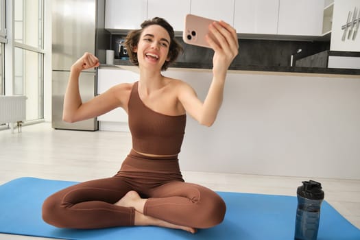 Portrait of young fitness woman, sitting at home on rubber mat, records video on smartphone, doing workout vlog, making live online training session from home. Copy space