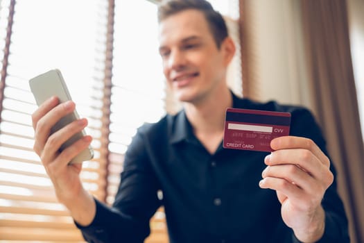 Young man sitting at table using online payment app and digital wallet on smartphone to pay with credit card. E commerce shopping and modern purchasing via mobile internet. Unveiling