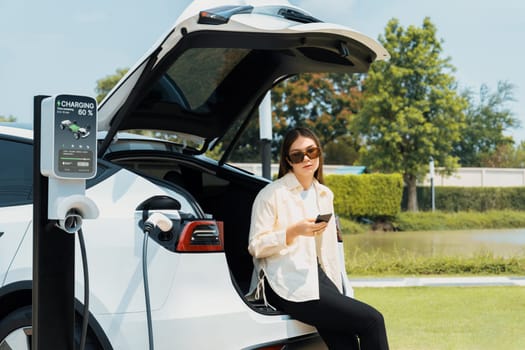 Young woman use smartphone to pay for electricity at public EV car charging station green city park. Modern environmental and sustainable urban lifestyle with EV vehicle. Expedient