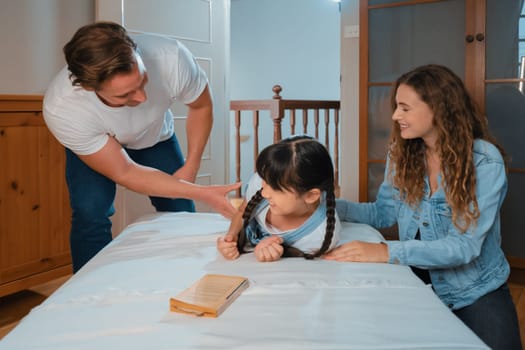 Happy modern family wakes up their little girl on the weekend with playful tickle expressing their love and affection for their young daughter, laughing and smiling together. Synchronos