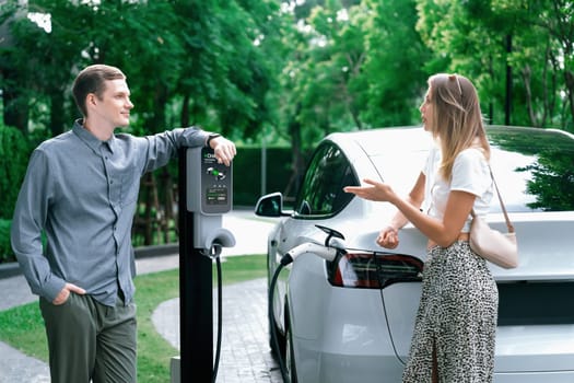 Young couple travel with EV electric car charging in green sustainable city outdoor garden in summer shows urban sustainability lifestyle by green clean rechargeable energy of electric vehicle innards