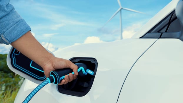 Hand insert EV charger and recharge electric car from charging station with wind turbine generator background. Smart eco-friendly EV car. Technological advancement of alternative energy. Peruse