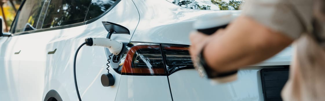 Young man recharge electric car's battery from charging station in outdoor green city park. Rechargeable EV car for sustainable environmental friendly urban travel. Panorama Expedient