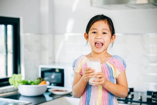 Adorable Asian preschooler holds milk cup in kitchen. Portrait of cute daughter enjoying drink smiling happily. Happy little girl relishing calcium-rich liquid radiating joy at home give me.