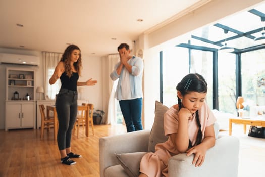 Annoyed and unhappy young girl sitting on sofa trapped in middle of tension by her parent argument in living room. Unhealthy domestic lifestyle and traumatic childhood develop to depression.Synchronos