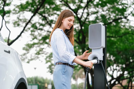 Young woman recharging battery for electric car during road trip travel EV car in natural forest or national park. Eco friendly travel during vacation and holiday. Exalt