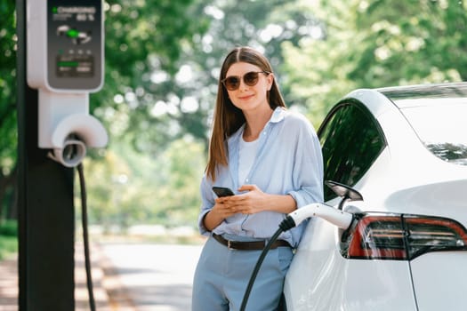 Young woman using smartphone online banking application to pay for electric car battery charging from EV charging station during vacation holiday road trip at national park or summer forest. Exalt