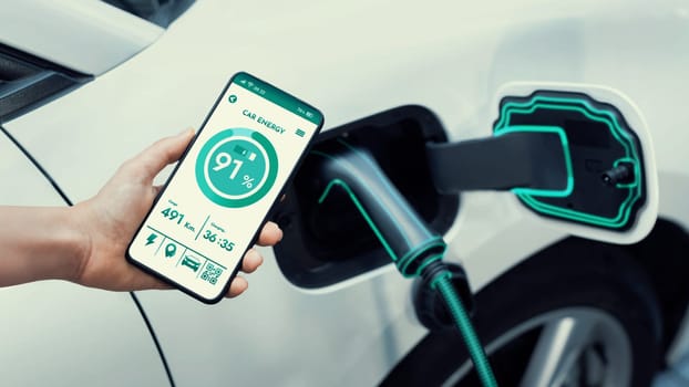 Businesswoman holding smartphone display battery status interface by smart EV mobile application while EV car recharging electricity from charging station in car park. Peruse