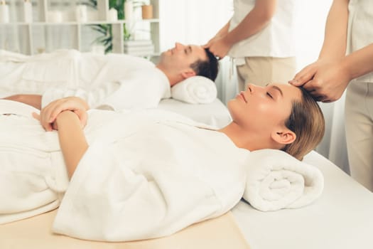Caucasian couple enjoying relaxing anti-stress head massage and pampering facial beauty skin recreation leisure in dayspa modern light ambient at luxury resort or hotel spa salon. Quiescent