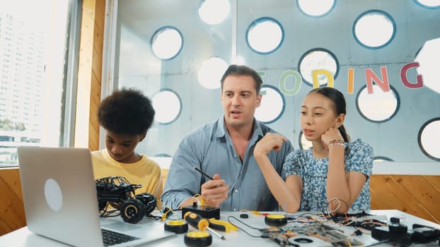 Caucasian student asking teacher about code and pointing at laptop screen. Professional instructor answer girl question while boy repair car model at table with electronic tool placed. Edification.