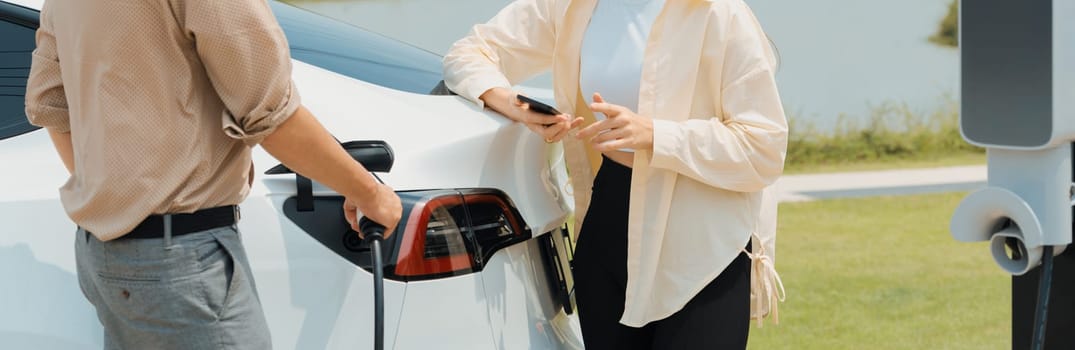 Young couple recharge electric car battery from charging station in green city park in springtime. Rechargeable EV car for sustainable environmental friendly urban travel lifestyle. Panorama Expedient