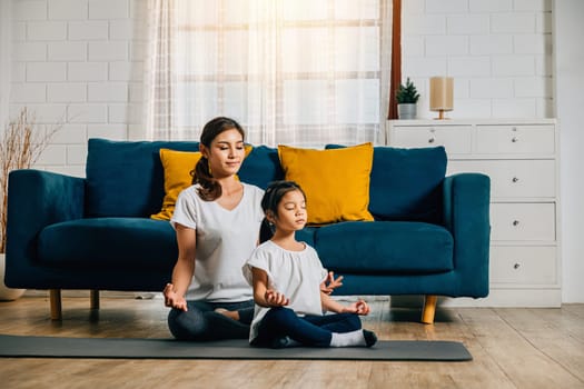Family yoga at home. A beautiful mother and her charming daughter smile while practicing yoga in lotus position promoting togetherness and mindfulness. Mom teaches her child to meditate.