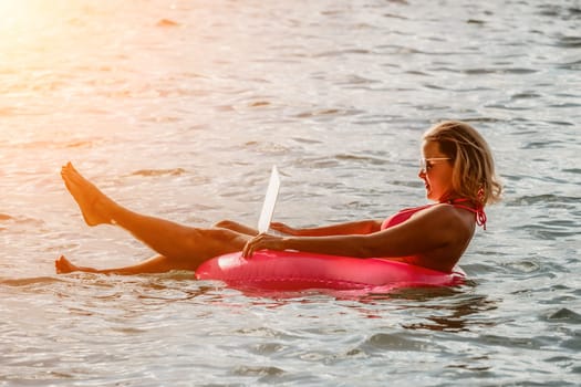 Woman works on laptop in sea. Freelancer, young blond woman in sunglases floating on an inflatable big pink donut with a laptop in the sea at sunset. Freelance, travel and holidays concept