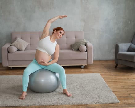 Pregnant red-haired woman doing arm exercises with fitball at home