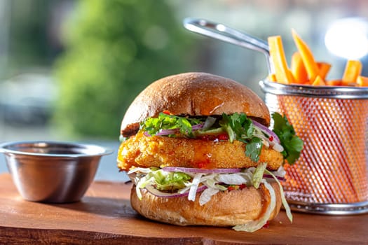 Hamburger with chicken cutlet, fresh tomatoes, red onion, lettuce and tomato sauce. Next to the burger is a metal mold with fries, a metal mold with chili sauce. Burger, fries and sauce are on a brown.