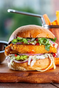 Hamburger with chicken cutlet, fresh tomatoes, red onion, lettuce and tomato sauce. Next to the burger is a metal mold with fries, a metal mold with chili sauce. Burger, fries and sauce are on a brown.