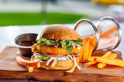 Hamburger with chicken cutlet, fresh tomatoes, red onion, lettuce and tomato sauce. Next to the burger is a metal mold with fries, a metal mold with chili sauce. Burger, fries and sauce are on a brown.