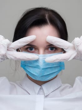 Young woman patient in a medical mask puts on protective surgical sterile gloves on her arm, on gray background, protection against coronovirus