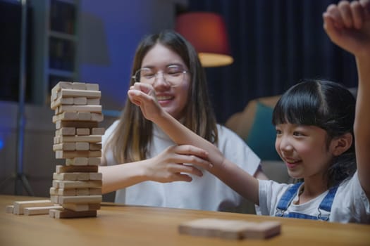 Asian young mother playing game in wood block with little daughter in home living room at night, Smiling woman help teach preschooler kid play build constructor tower of wooden blocks, family funny