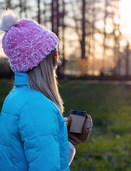 A beautiful cute girl in a hat and a down jacket holds a paper cup with hot coffee or tea. Frosty sunny morning Winter season.