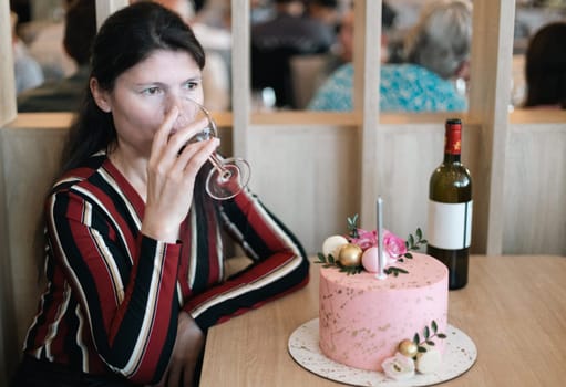 One beautiful Caucasian brunette girl sits at a table in a restaurant with a pink cake and looking to the side drinks red wine from a glass celebrating her birthday, side view close-up with depth of field.