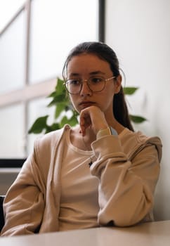 Portrait of one beautiful caucasian brunette teenager girl in glasses in beige clothes sits in a cafe against the background of a panoramic window looking to the side and holding her chin with her hand, side view close-up.
