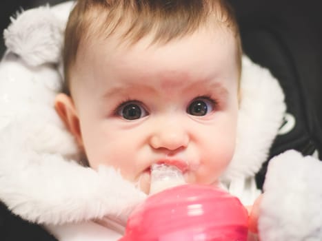 Portrait of one beautiful Caucasian baby girl with brown eyes drinking milk from a bottle with a pacifier while sitting in a stroller in a cafe on a blurred background, close-up side view with depth of field.