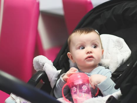 Portrait of one beautiful Caucasian girl with brown eyes holding a pink bottle with pacifier and milk in her hands, sitting in a black stroller in a cafe and looking carefully to the side, on a blurred background, close-up side view with depth of field.