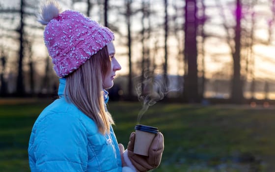 A beautiful cute girl in a hat and a down jacket holds a paper cup with hot coffee or tea. Frosty sunny morning Winter season.