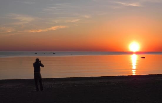 Back view of a silhouette of a man photographing the sun at sunset on the beach on holidays.