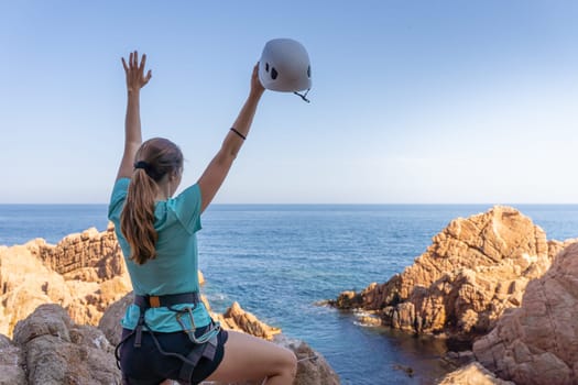 Pretty female climber celebrating strong success with arms raised on rocks over the sea with rope and helmet. High quality photo