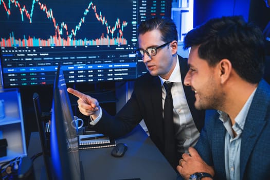 Two stock exchange traders discussing on dynamic successful investment graph, pointing trending of high profit in currency stock in financial data on monitor in neon light at workplace. Sellable.