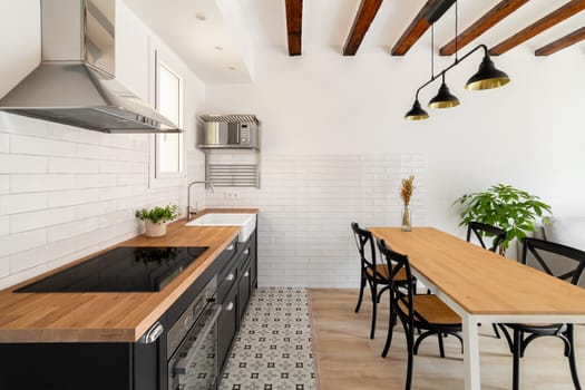 Long dining table with four chairs next to a kitchen island with a build in stove and extractor hood with white walls and renovation beams in a newly built house. Copyspace.