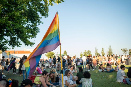 Lisbon, Portugal. 17 June 2023: Picnic in park in Lisbon during Gay Pride Parade. Rainbow flag flying over people sitting on grass. Happy and cheerful people on lgbtqia pride event