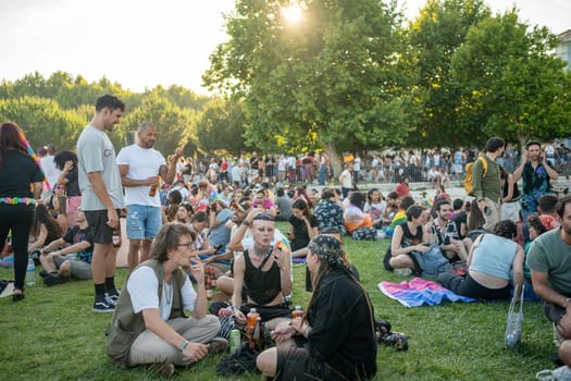 Lisbon, Portugal. 17 June 2023 Activists preparing for Pride Parade in park. People sitting on grass surrounded by LGBTQ posters, rainbow flags. People using cell phones in expectation of Pride Parade