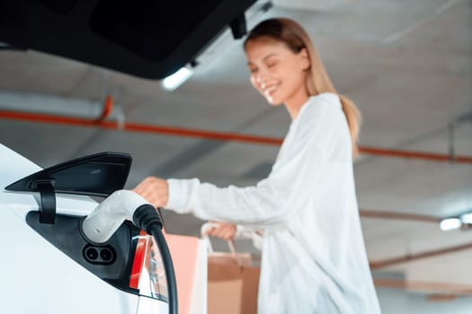 Young woman travel with EV electric car to shopping center parking lot charging in downtown city showing urban sustainability lifestyle by green clean rechargeable energy of electric vehicle innards