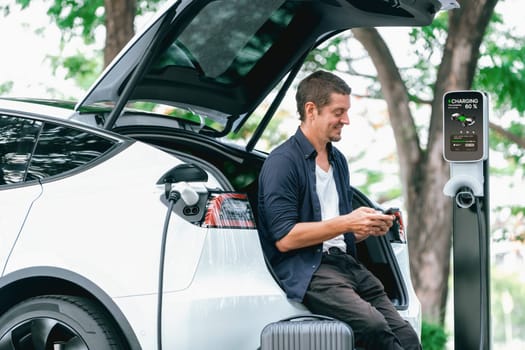 Man using smartphone online banking application to pay for electric car battery charging from EV charging station during vacation holiday road trip at national park or summer forest. Exalt