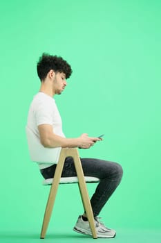 A man, on a green background, is sitting on a chair, with phone.