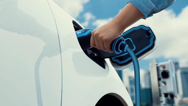 Asian man insert EV charger and recharge his electric car from future charging station with blurred background of cityscape. Smart and futuristic sustainable energy infrastructure. Peruse