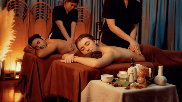 Caucasian couple customer enjoying relaxing anti-stress spa massage and pampering with beauty skin recreation leisure in warm candle lighting ambient salon spa at luxury resort or hotel. Quiescent