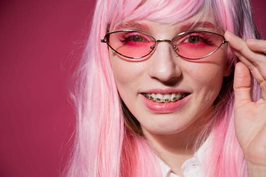 Close-up portrait of a young woman with braces in a pink wig and sunglasses on a pink background