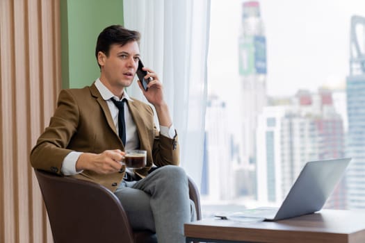 Closeup of handsome businessman making phone call with manager while sitting near window with skyscraper view. Executive manager talking working by using phone and laptop. Look aside. Ornamented.