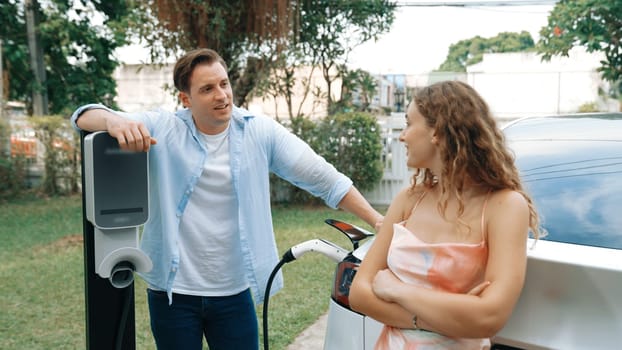 Happy and lovely couple with eco-friendly conscious recharging electric vehicle from EV charging station. EV car technology utilized as alternative transportation for future sustainability. Synchronos