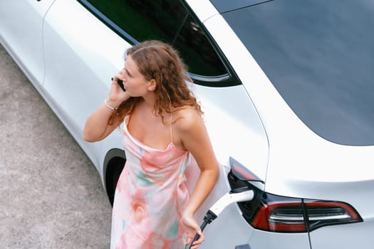 Aerial top view eco-friendly woman recharge electric vehicle from EV charging station, using EV technology utilization for tracking energy usage to optimize battery charging on smartphone. Synchronos