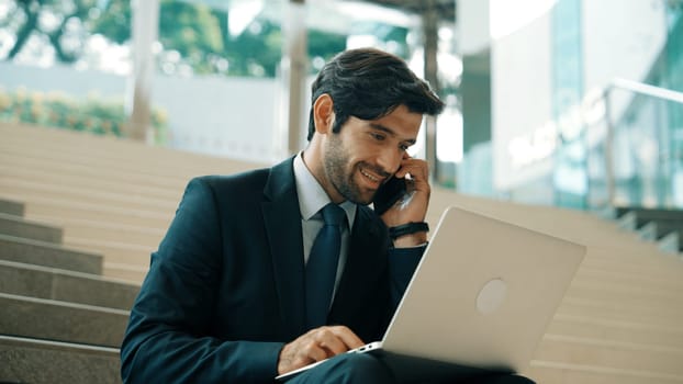 Skilled investor working or planing strategy by using laptop at stair. Professional business man wearing suit while working and typing data analysis by using laptop and talking to phone. Exultant.